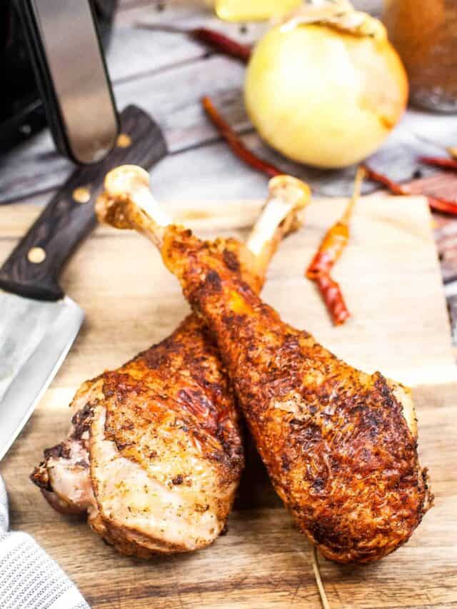Two chicken legs on a cutting board next to an air fryer.