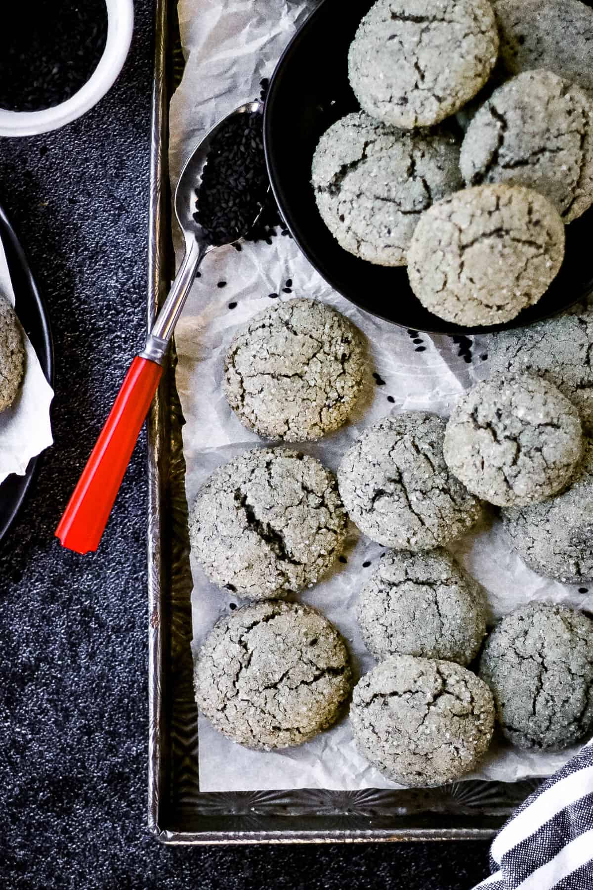overhead shot of black sesame cookies on a baking sheet with parchment paper and a red-handled spoon.
