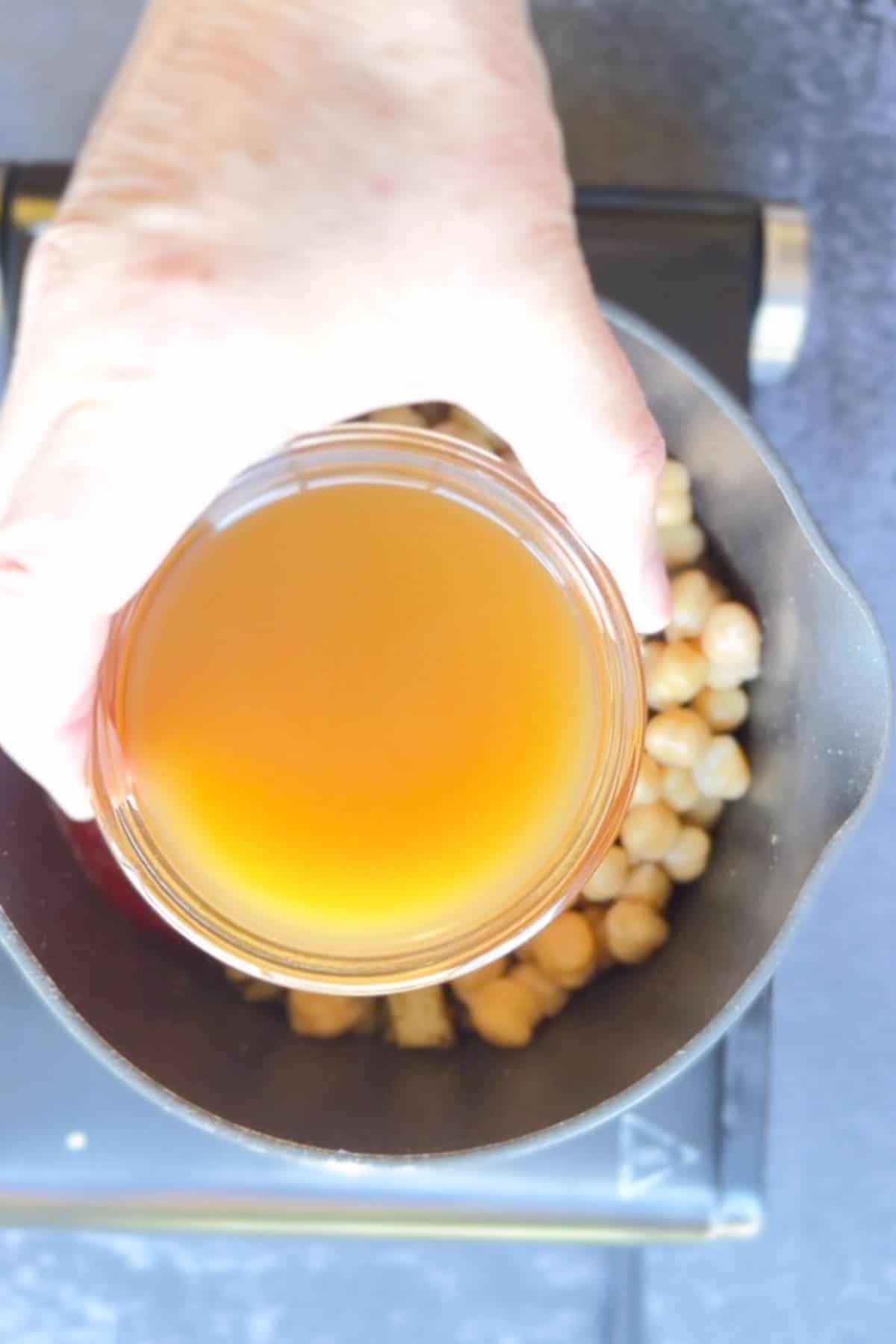A person pouring broth into a pan with chickpeas.