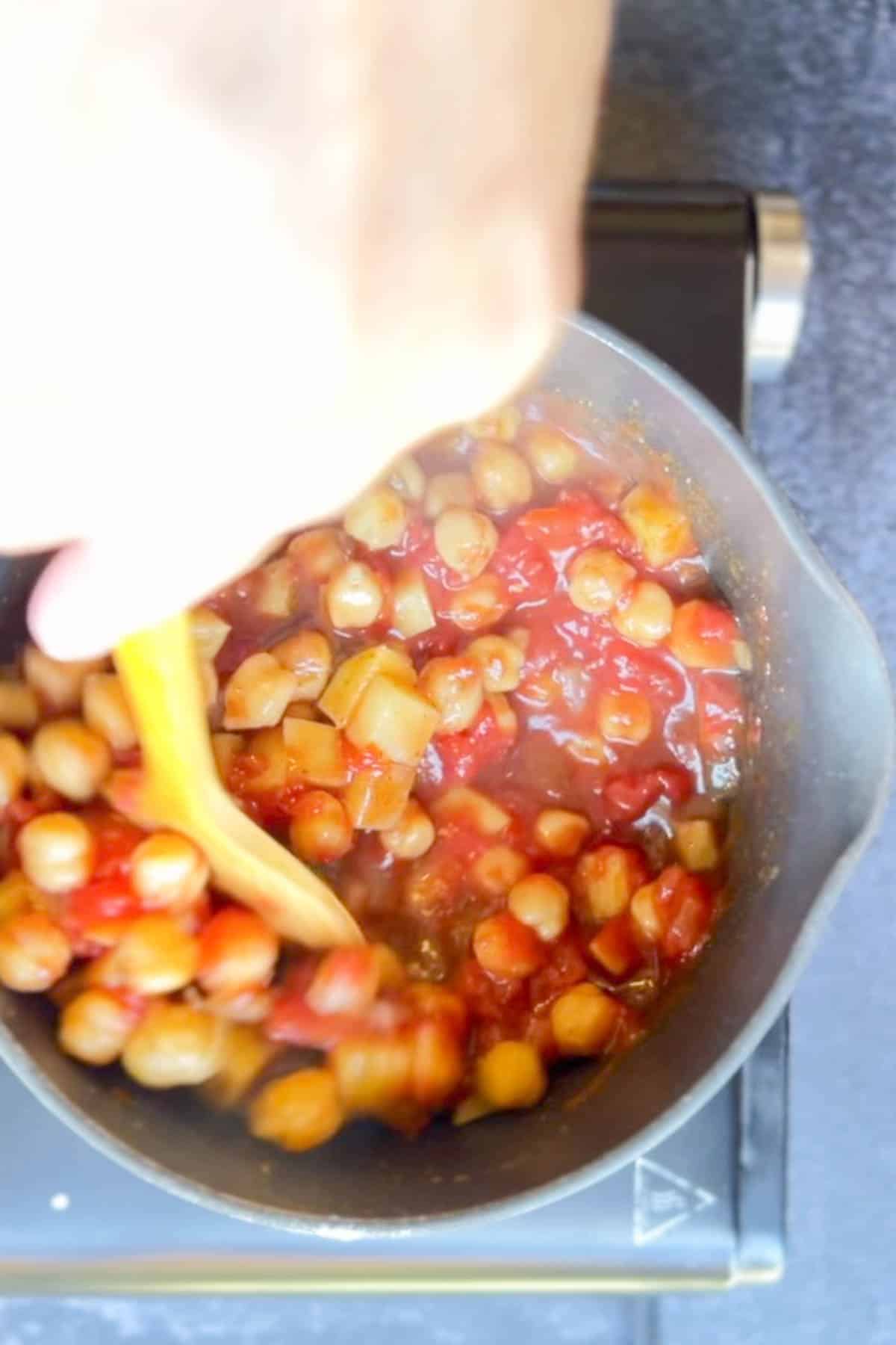 A person cooking chana aloo masala in a pan.