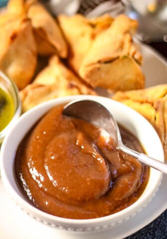 low angle shot of a bowl of tamarind chutney with a spoon and samosas in the background.