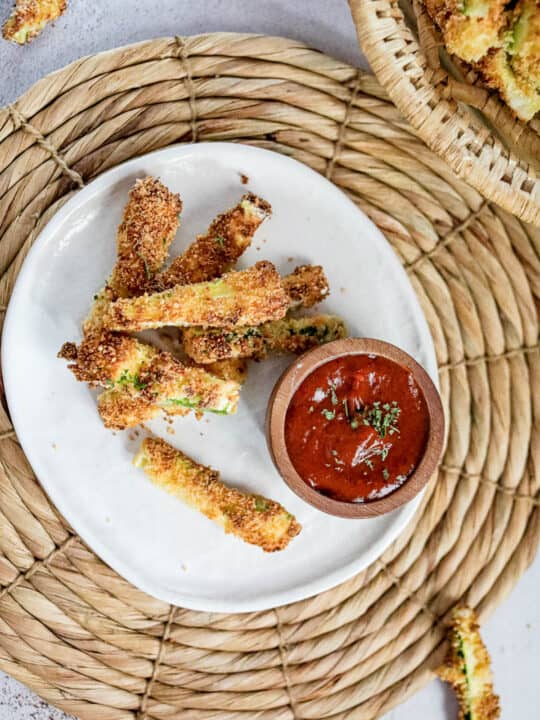 Overhead shot of air fryer zucchini fries on a white plate with a small bowl of spicy ketchup dipping sauce.