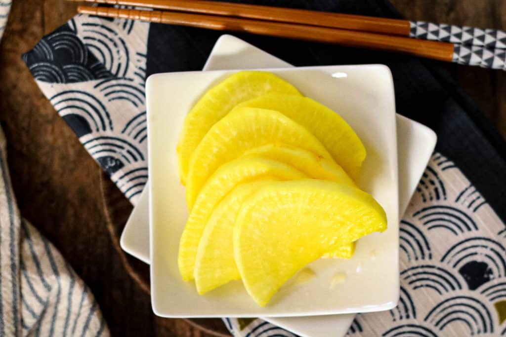 A white square plate with several slices of yellow pickled daikon radish, placed on a patterned cloth next to wooden chopsticks.