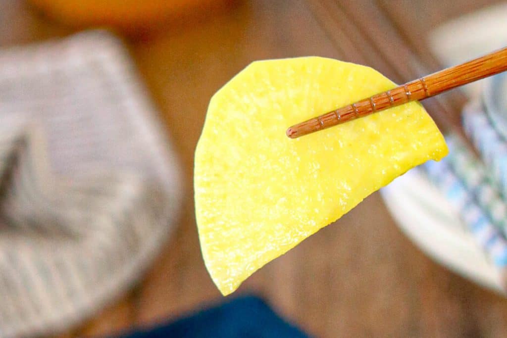 A pair of chopsticks holding a single slice of pickled daikon radish against a blurred background.