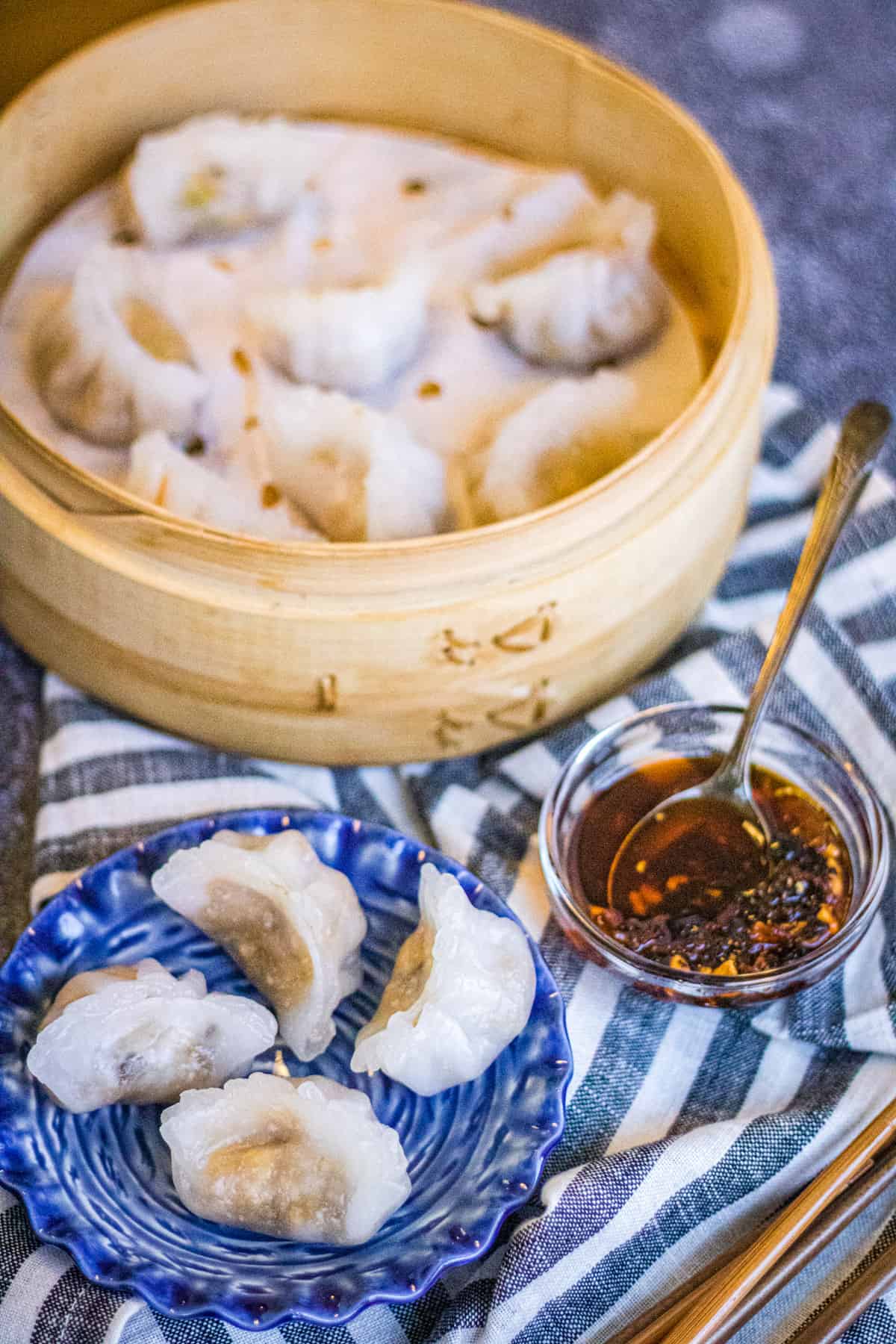 low angle shot of crystal vegetarian dumplings on a blue plate with a steamer basket of dumplings and a bowl of chili crisp dipping sauce on the side.