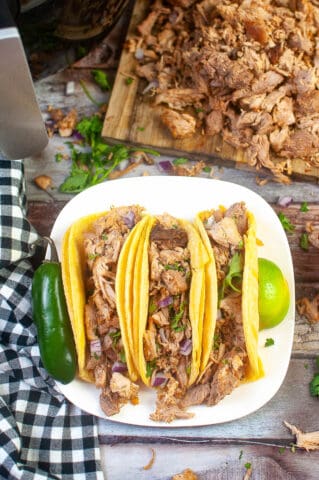 High angle shot of 3 carnitas tacos on a plate with more carnitas on a cutting board in the background.