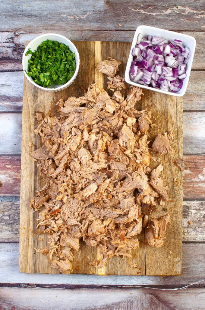 Step five for making air fryer carnitas. The cooked meat shredded on a cutting board with cilantro and diced red onion.