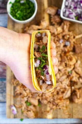 Overhead shot of a hand holding a taco stuffed with carnitas. There is a cutting board covered with carnitas in the background.