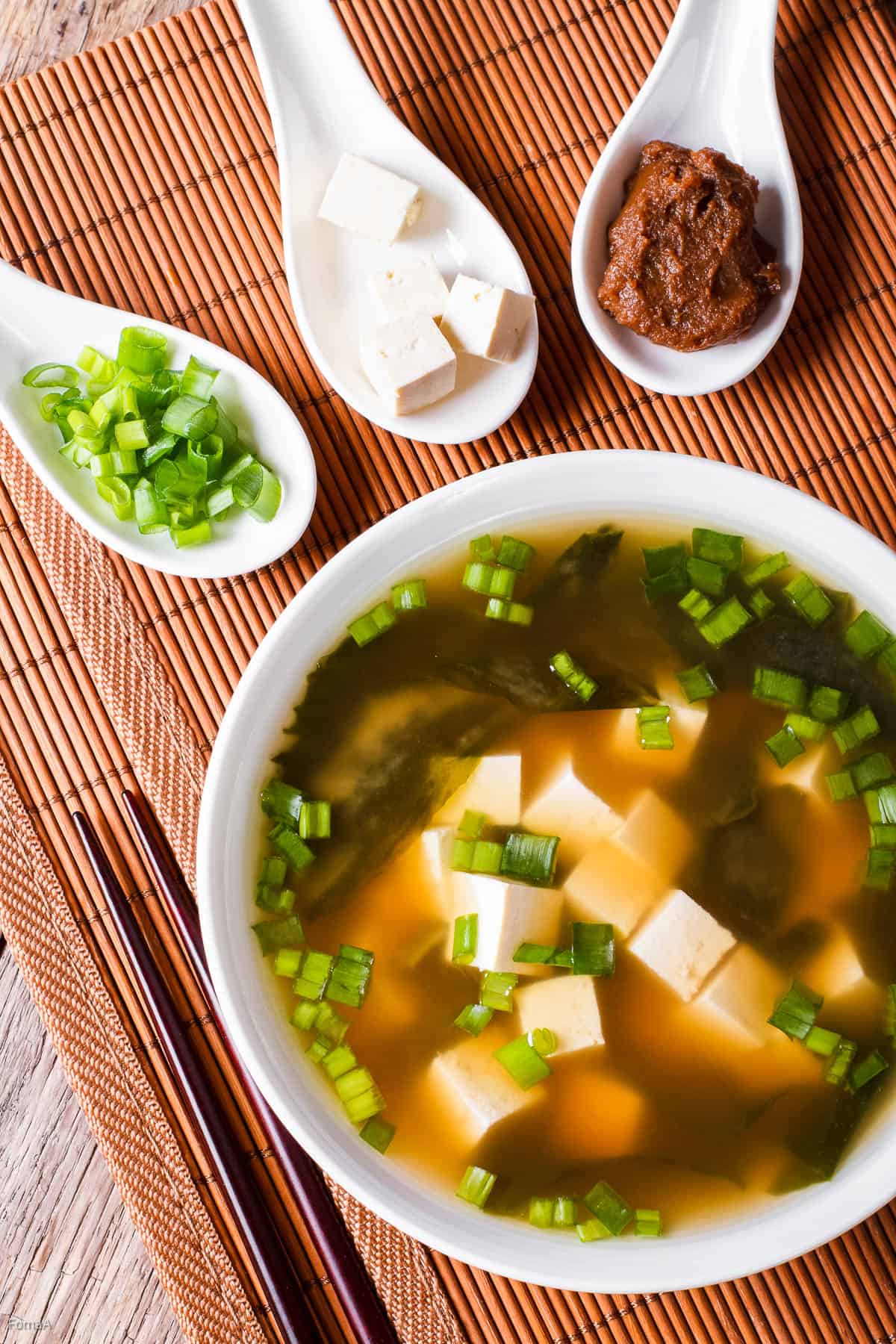 Overhead shot of a bowl of miso soup. There are 3 Asian style soup spoons on the side filled with green onions, tofu cubes, and miso paste.