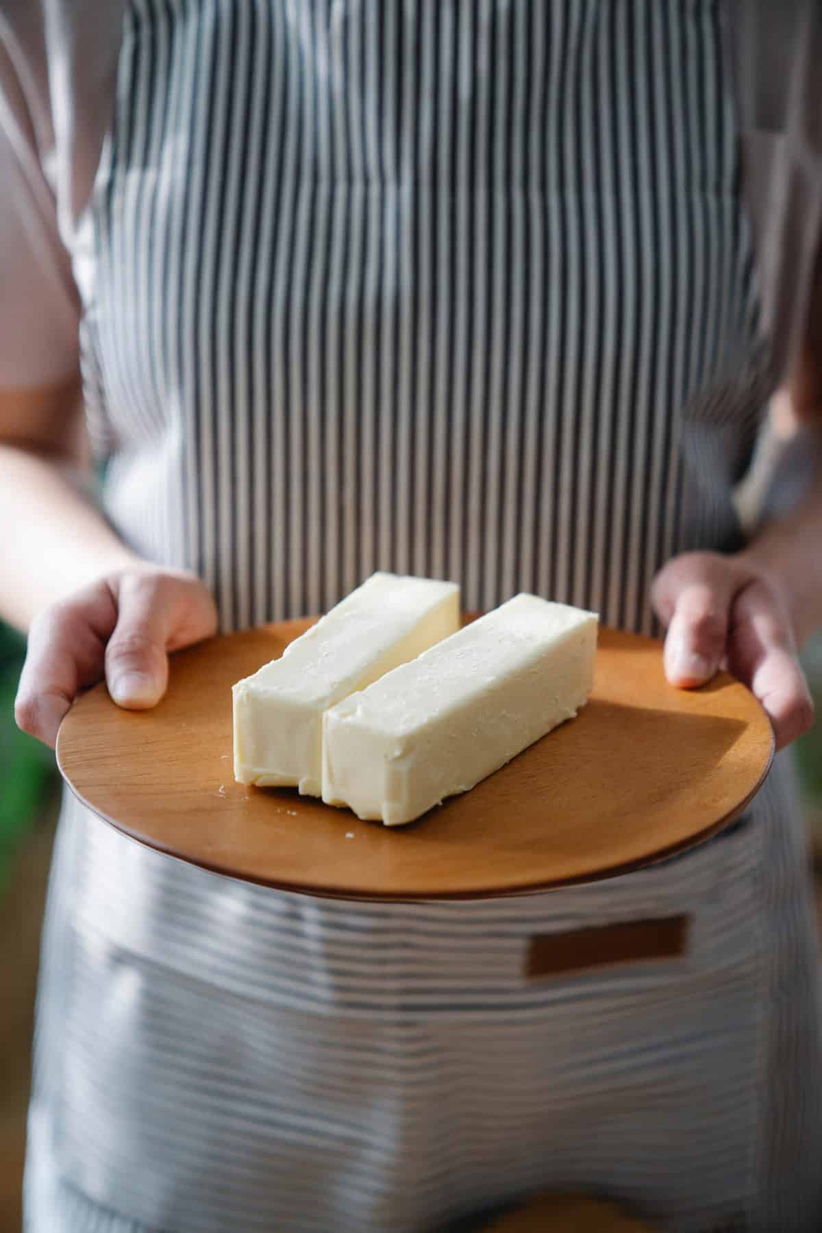 A person holding a wooden plate with two sticks of softened butter on it. You can only see the persons torso  in a striped apron and their hands. 