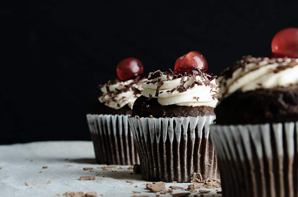 Low angle shot of 3 chocolate cupcakes with buttercream frosting and maraschino cherries.