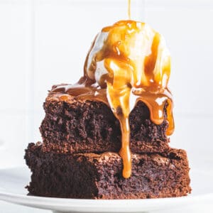 2 brownies stacked on a plate with a scoop of vanilla ice cream on top drizzled with caramel.