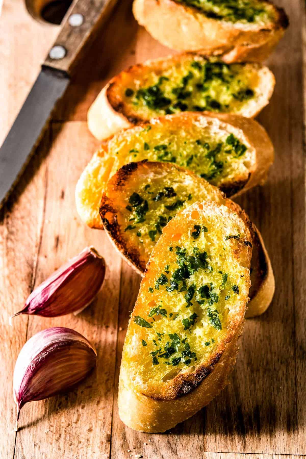 Slices of garlic bread on a wooden board with a knife and a couple of cloves of purple garlic.