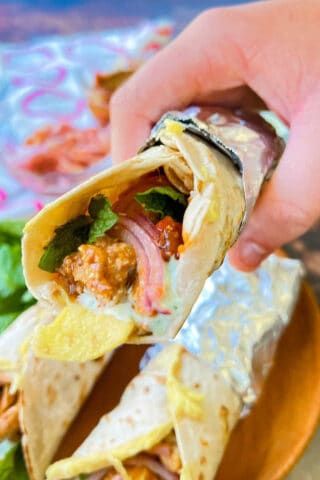 A chicken kathi roll wrapped in foil with a hand holding it.