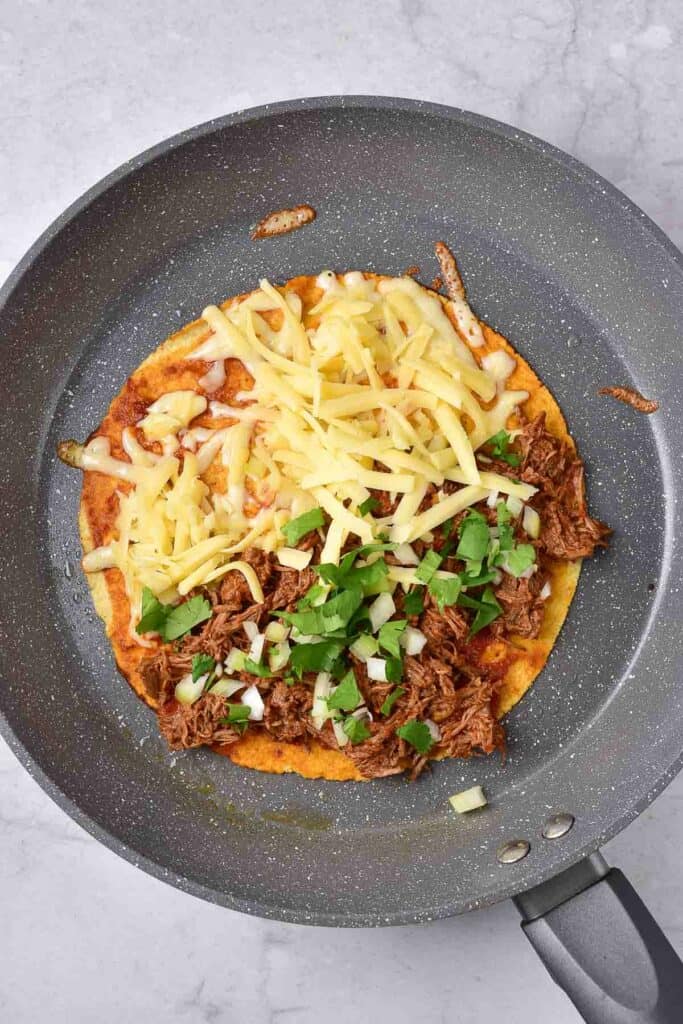 tortilla with birria, cheese, cilantro and onion in a skillet.