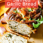 A loaf of swirled garlic bread with herbs, partially sliced, sits on a wooden cutting board alongside fresh garlic cloves, ready to enjoy.