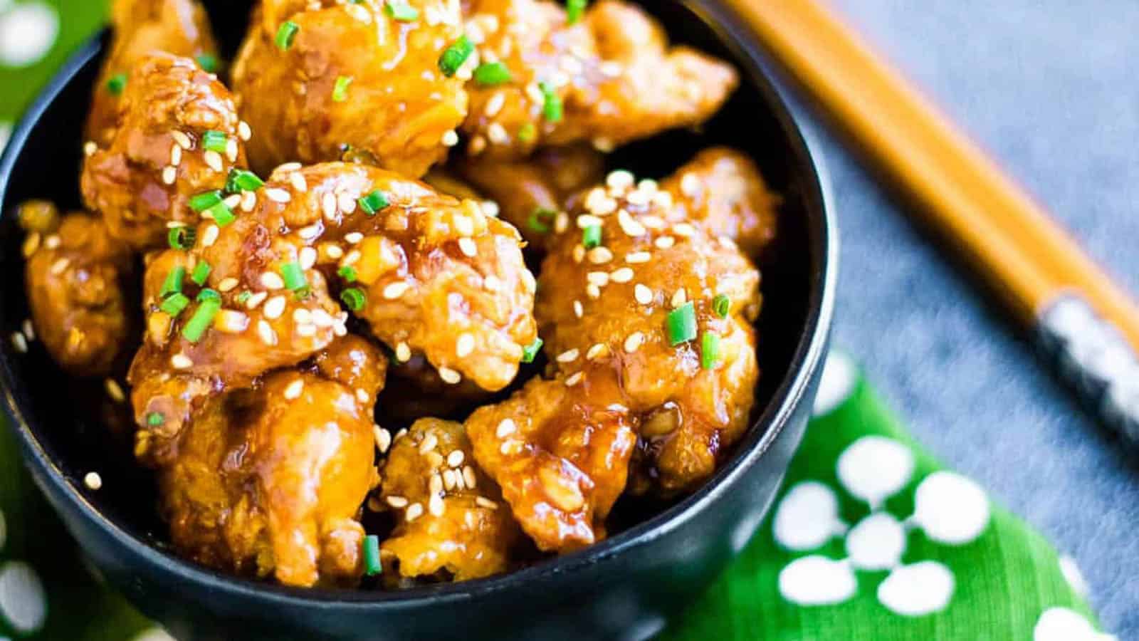 Fried chicken pieces in orange sauce garnished with sesame seeds in a black bowl with chopsticks and a green and white napkin.