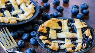 Two small blueberry pies with lattice crusts sit on a wooden surface, surrounded by fresh blueberries and two forks—perfect for enjoying decadent desserts with a touch of homemade charm.