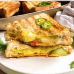 Two grilled Bombay sandwiches filled with vegetables and melted cheese are stacked on a white plate. The top sandwich is garnished with sliced green chilies, adding a spicy kick. In the background, a baking dish brims with more delectable Bombay sandwiches, ready to be enjoyed.