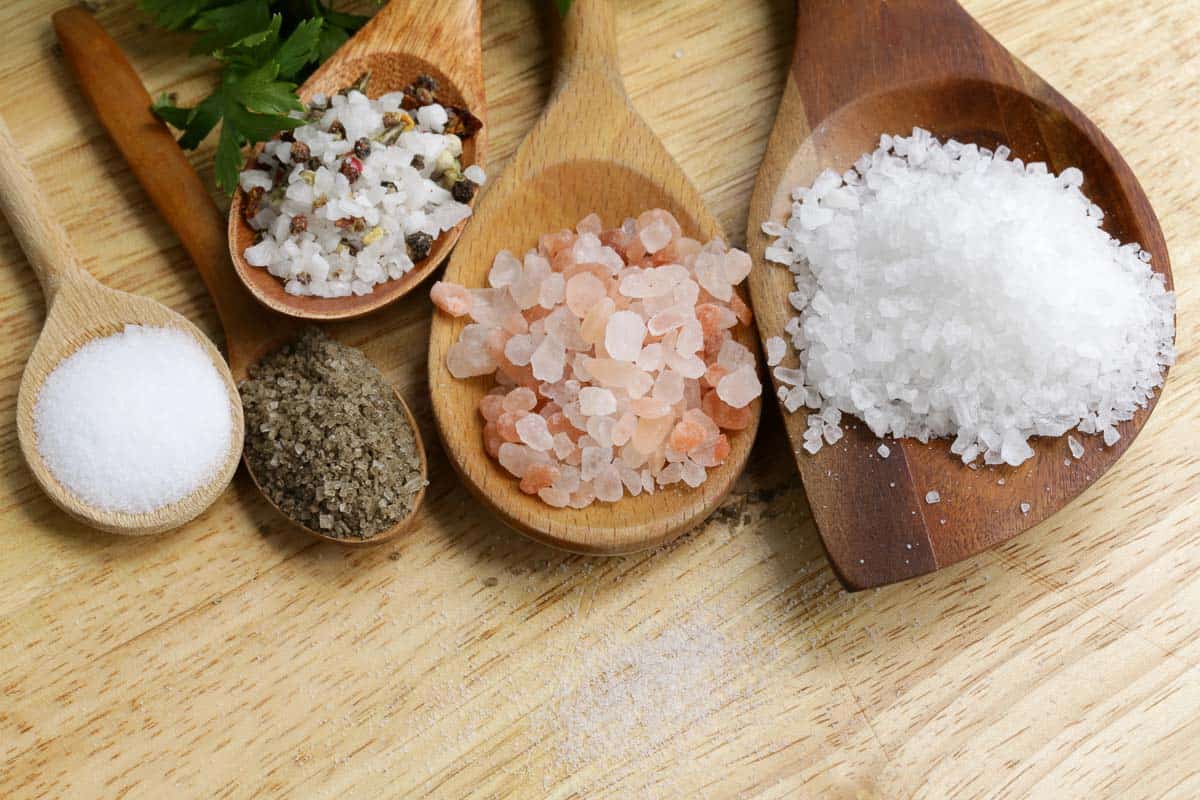 Different types of salts and herbs arranged on wooden spoons, resting on a rustic wooden table.