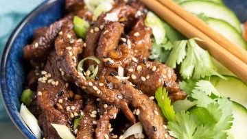 A bowl of sesame beef stir-fry with chopsticks and fresh cucumber slices on the side.