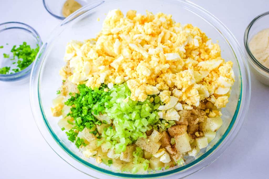 A bowl of potato salad ingredients, including chopped boiled eggs, diced potatoes, celery, and herbs, in the process of being mixed.