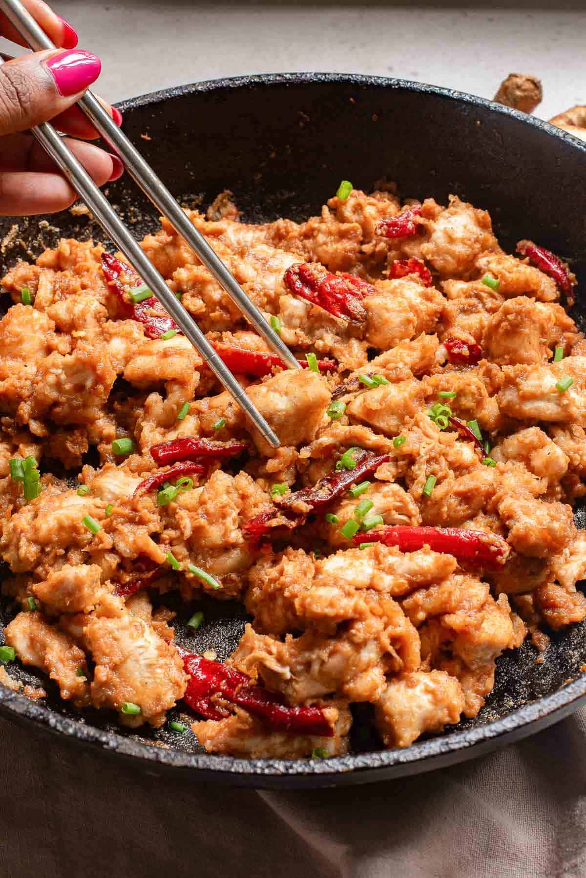Stir-frying chicken with red peppers in a black skillet using metal tongs.