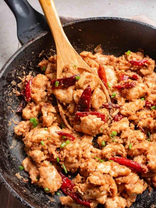 Stir-fried chicken with chili peppers and herbs in a black pan with a wooden spatula.