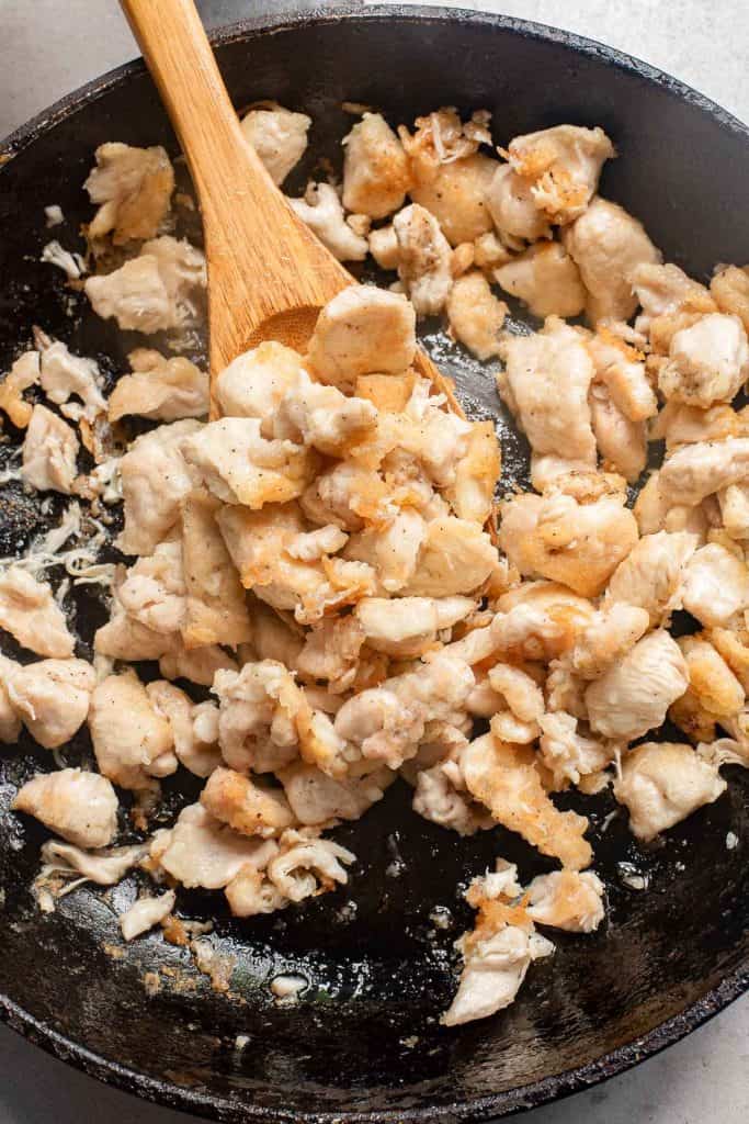 Cooked chicken pieces being stirred in a black frying pan with a wooden spatula.