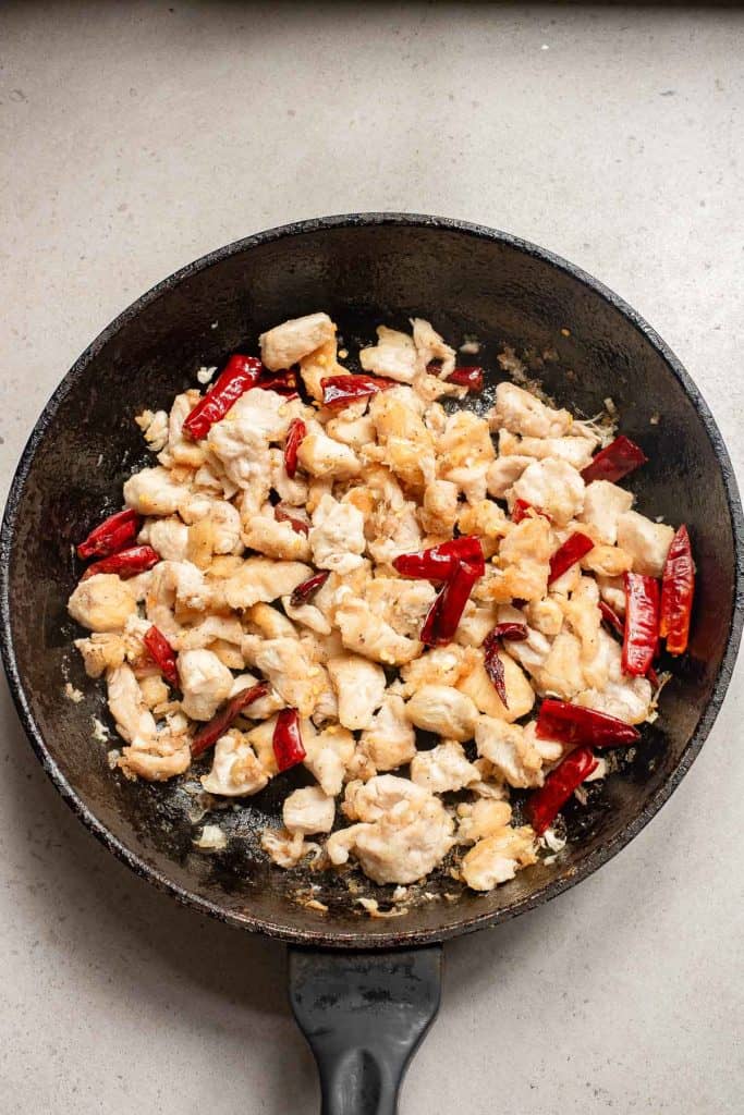 Chicken pieces being stir-fried with red chili peppers in a skillet.