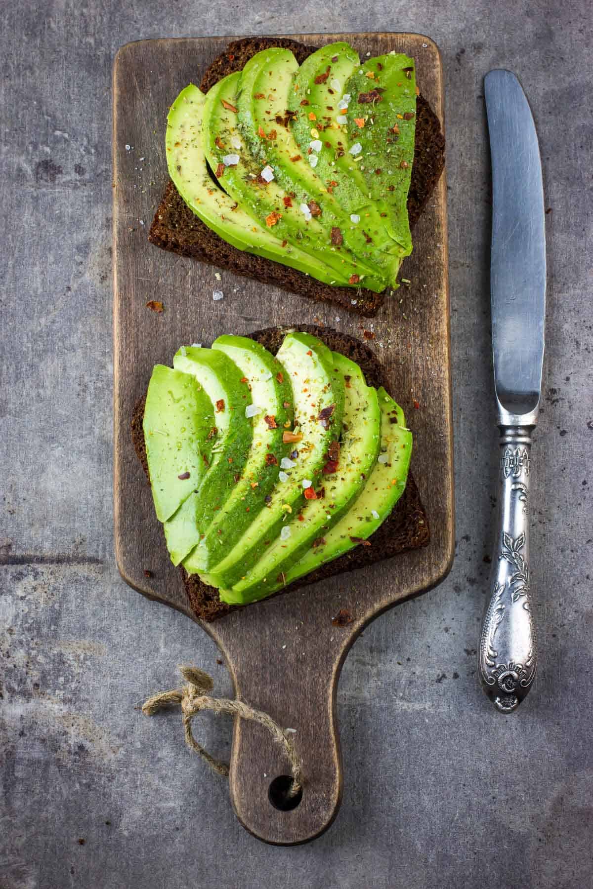 Two slices of bread topped with avocado slices, sprinkled with seasoning, on a wooden cutting board next to a knife. By the way, can you freeze avocados to keep your favorite toppings on hand longer?