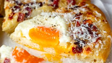 Close-up of a carbonara pizza slice topped with a runny egg, grated cheese, and bits of bacon.