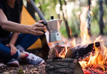 A person holding a moka pot over a campfire next to a yellow tent in a forest setting.