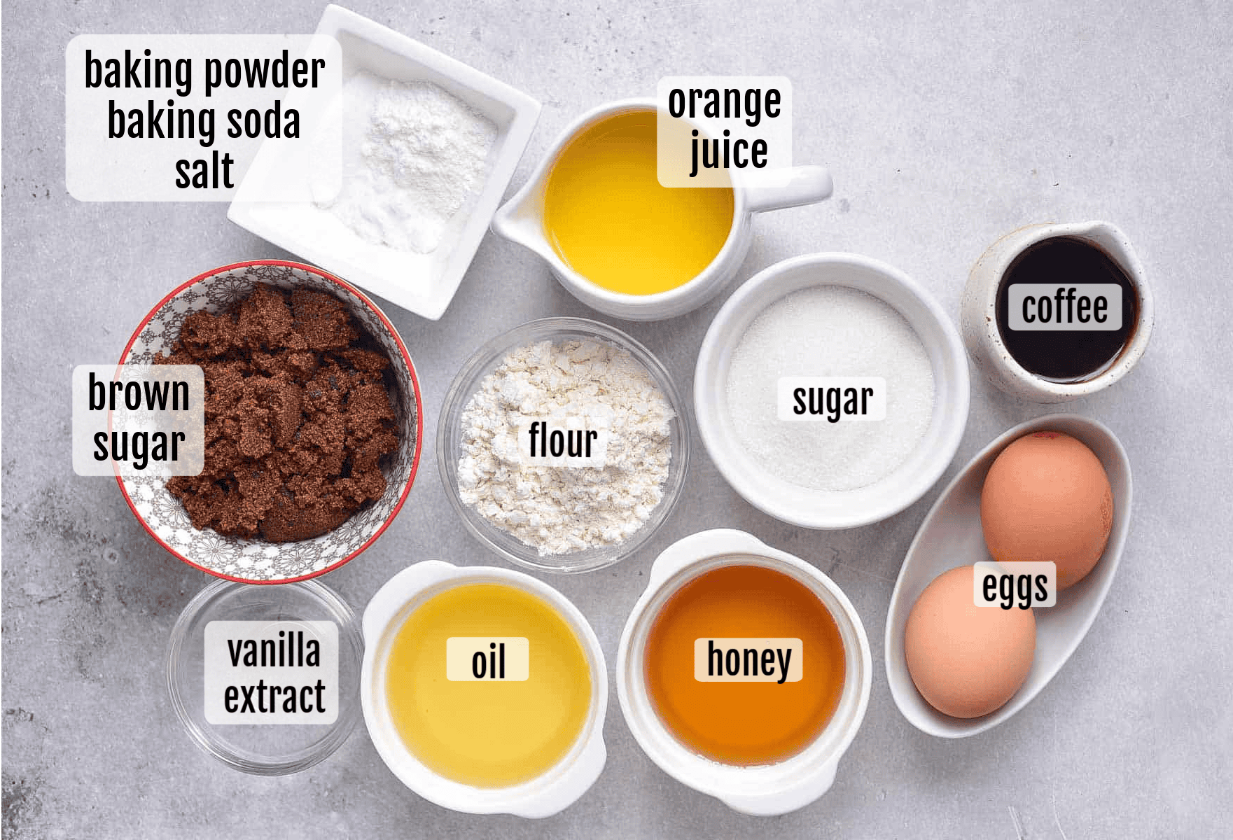 Various labeled baking ingredients for honey cake in bowls and measuring cups, including brown sugar, flour, eggs, oil, honey, vanilla extract, coffee, orange juice, sugar, baking powder, baking soda, and salt.