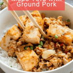 A bowl of white rice topped with cubes of tofu, ground pork in sauce, chopped green onions, and two wooden chopsticks.