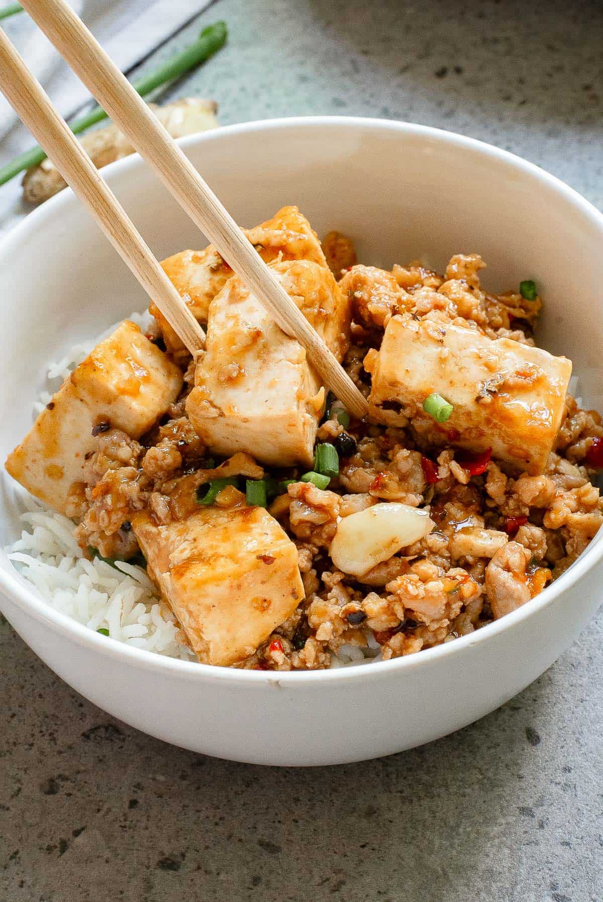 A bowl of tofu stir-fry with vegetables and sauce is served over a bed of rice. Chopsticks are placed in the bowl.