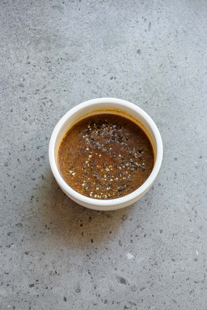 A white bowl filled with dark brown liquid sauce speckled with black and white seeds sits on a gray textured surface.