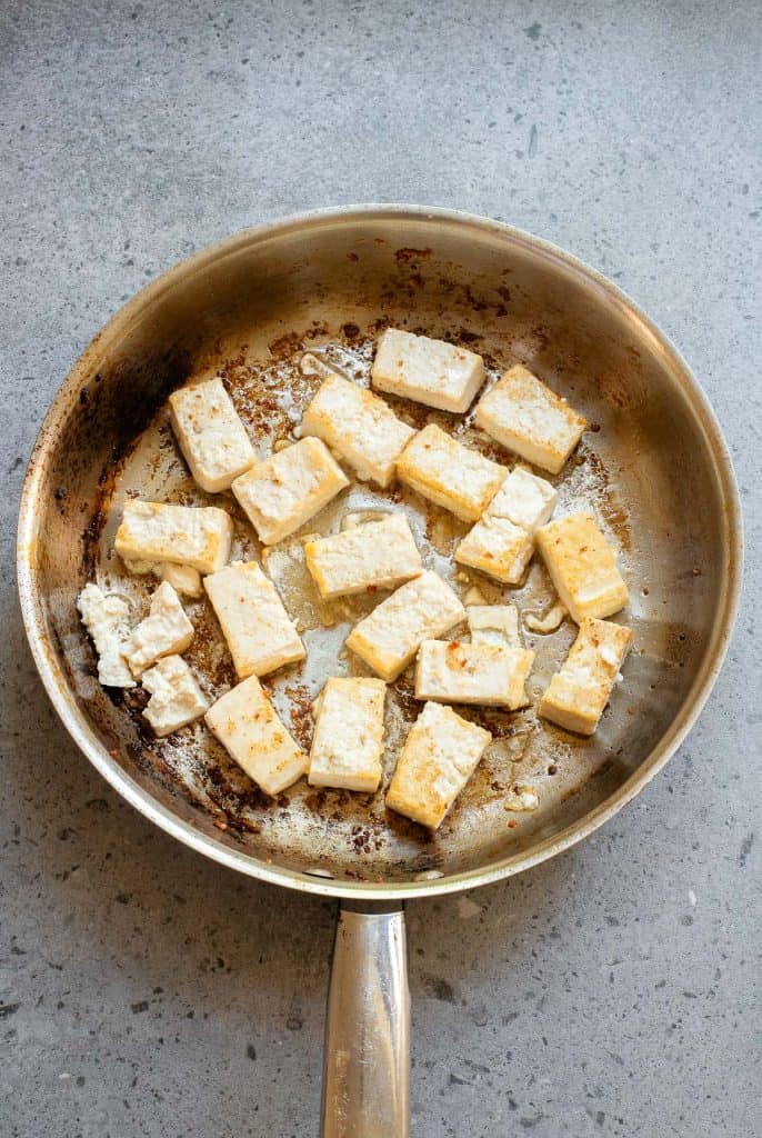 A frying pan containing browned cubes of tofu on a grey countertop.