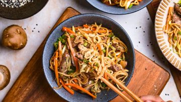 A bowl of stir-fried noodles with vegetables and beef, garnished with sesame seeds. Chopsticks are used to pick up the noodles. Other dishes and ingredients are partially visible around it.