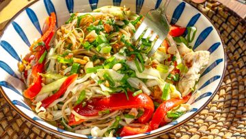 A bowl of colorful noodle salad with red bell peppers, shredded cabbage, chopped green onions, and crushed peanuts. A fork is placed within the salad.