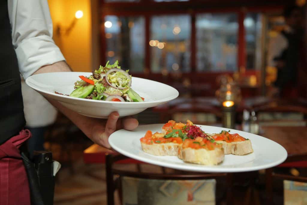 A server holding two plates of food: one with a mixed green salad and the other with bruschetta topped with diced tomatoes and herbs in a restaurant setting.