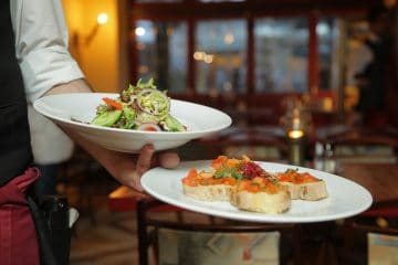 A server holding two plates of food: one with a mixed green salad and the other with bruschetta topped with diced tomatoes and herbs in a restaurant setting.
