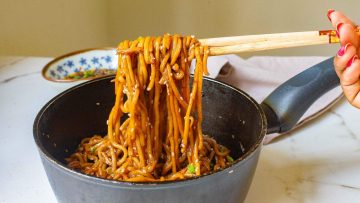 A close-up of a hand using chopsticks to lift cooked noodles from a black pan. The noodles are mixed with vegetables and sauce. A small dish is visible in the background.