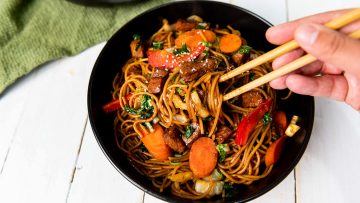 A bowl of stir-fried noodles with beef, carrots, and bell peppers. A hand uses chopsticks to pick up noodles.