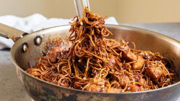 A frying pan filled with stir-fried noodles and chunks of meat, being tossed with chopsticks.
