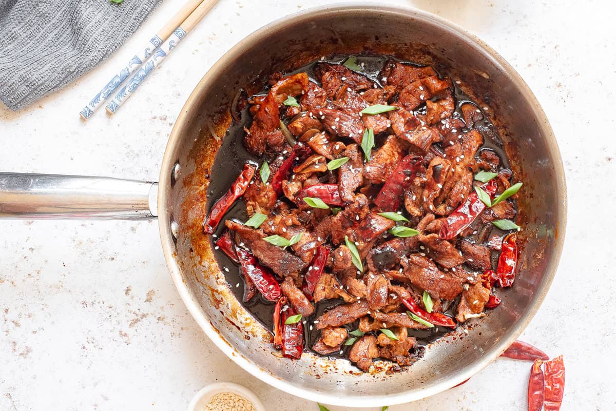 A pan of spicy beef stir-fry with red chili peppers and green onions on a white surface, surrounded by a napkin, chopsticks, and a small bowl of sesame seeds.