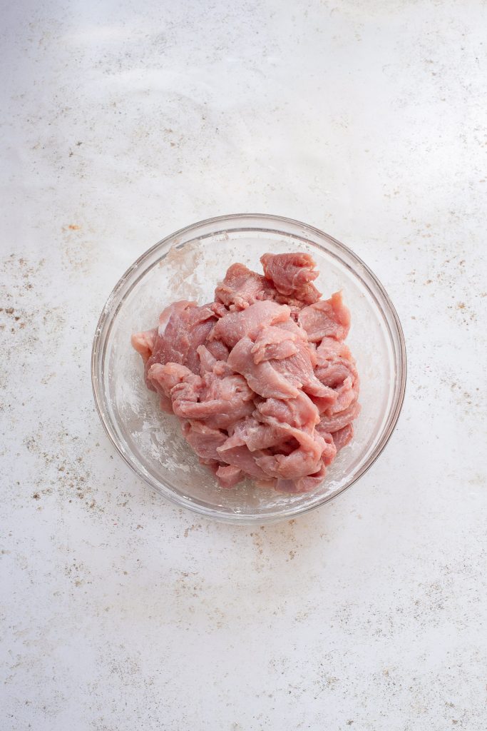 A clear glass bowl filled with raw, sliced pork pieces on a white, textured surface.