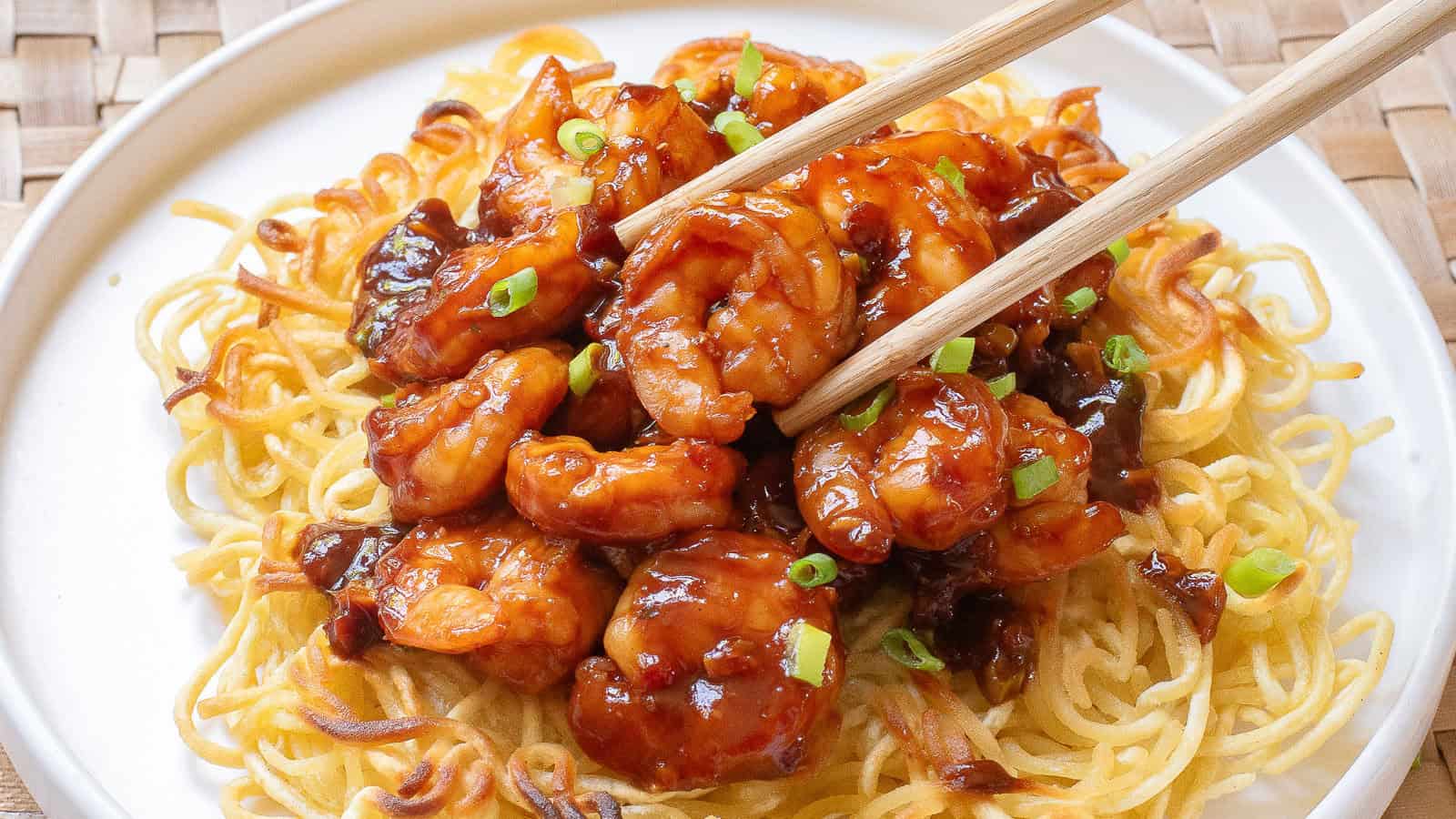 Chopsticks holding a spicy prawn on a bed of crispy noodles, garnished with green onions, served on a white plate.