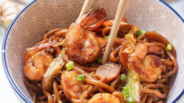 Bowl of stir-fried noodles with shrimp, vegetables, and chopsticks.