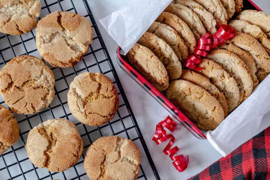 Cookies on a cooling rack and in a plaid tin lined with parchment paper, with red ribbon decorations nearby.